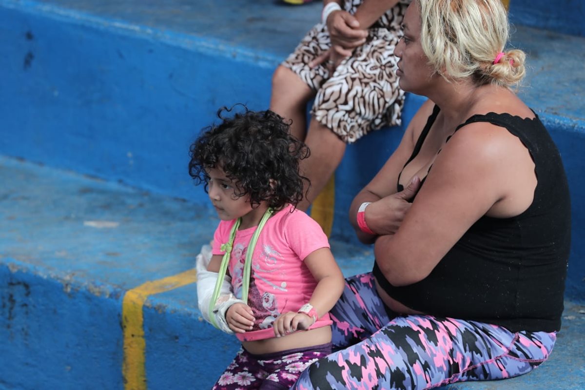 Una niña con su brazo quebrado espera por medicina mientras otro familiar hace la fila para recibirla.