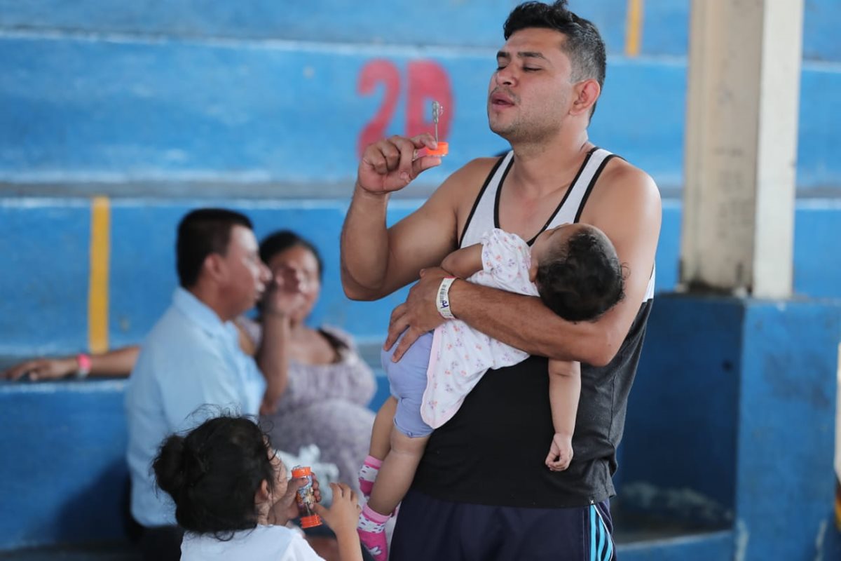 Un padre distrae a su bebé con burbujas mientras hace fila para recibir medicinas.