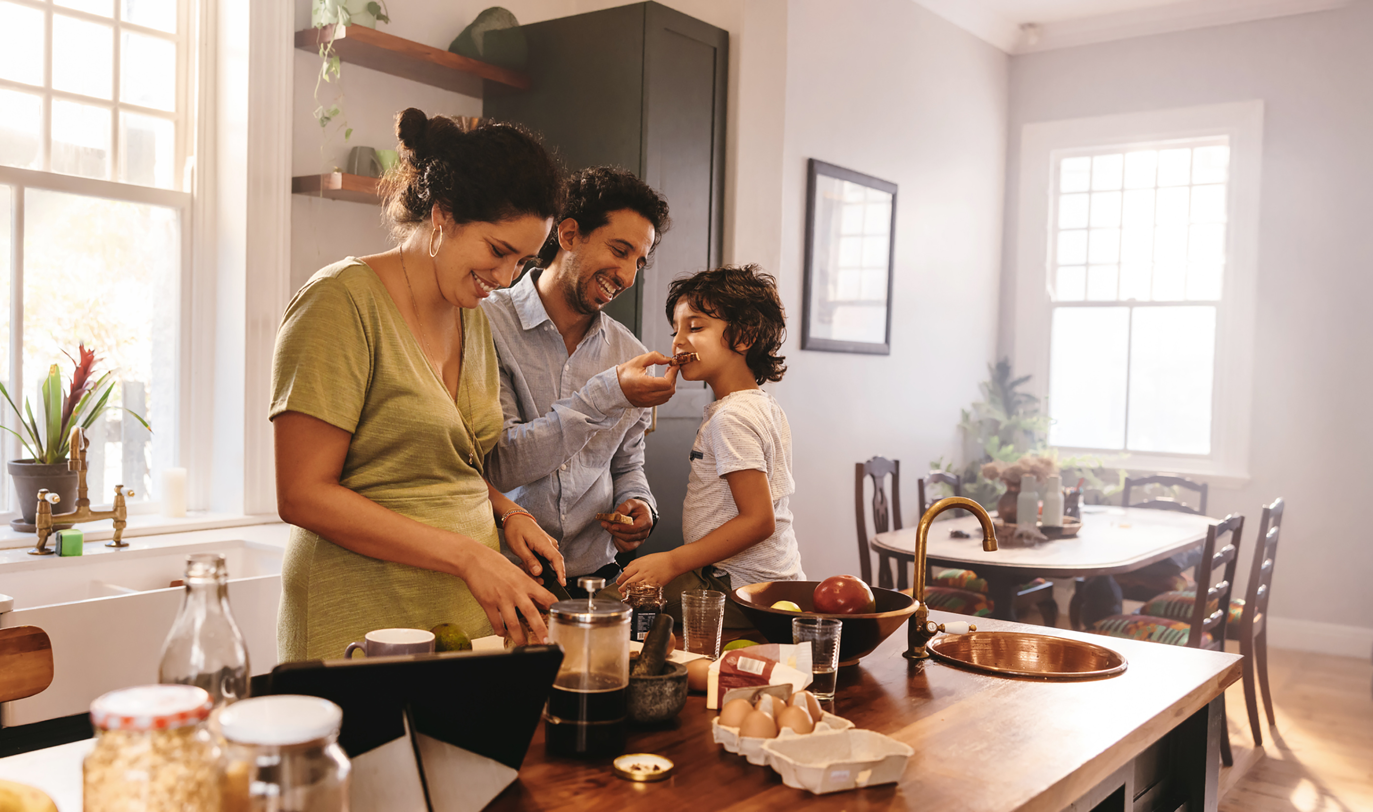 Playful dad feeding his son a slice of bread while his wife prepares breakfast. Family of three having fun together in the kitchen. Mom and dad spending quality time with their son.