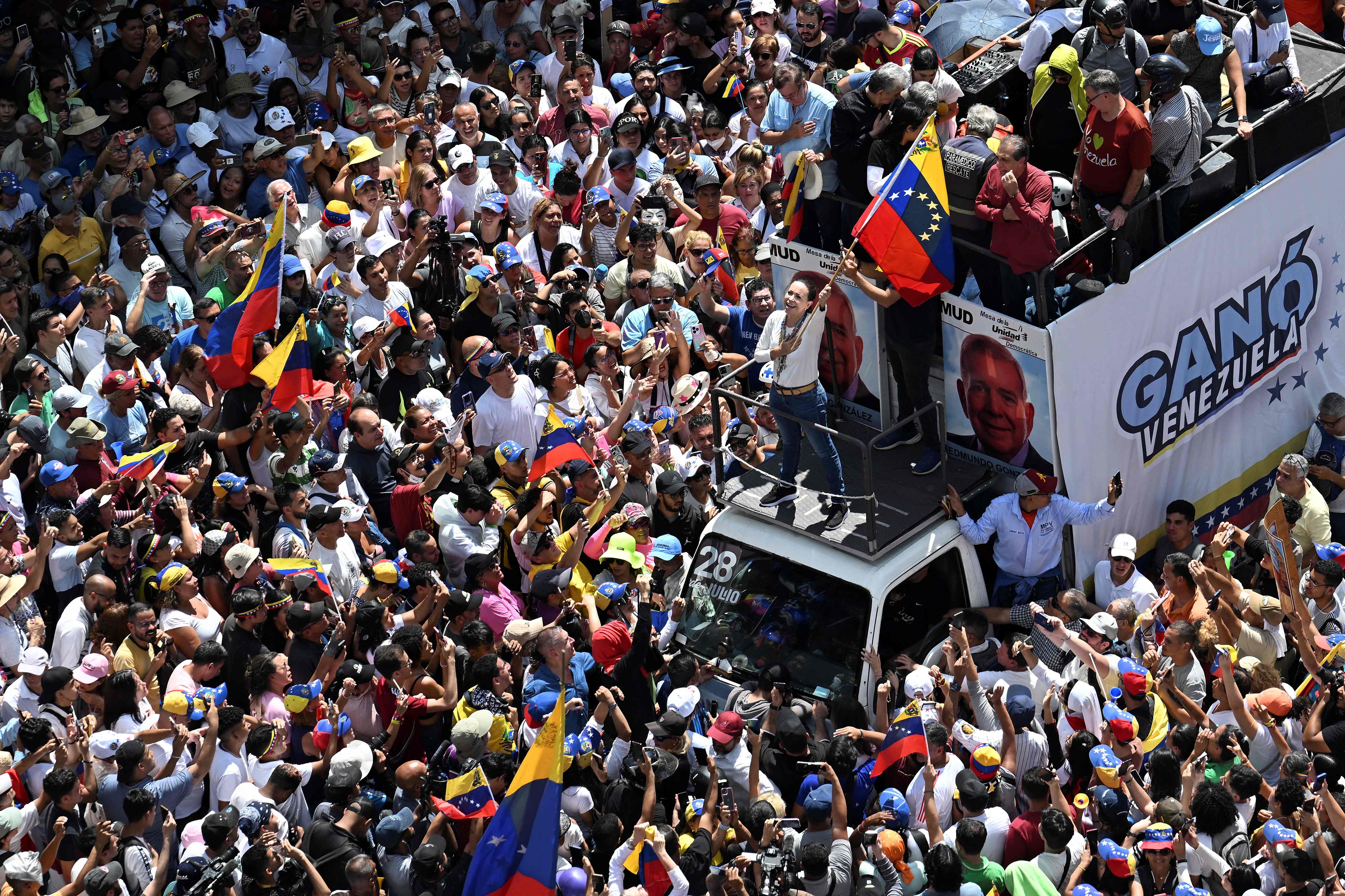 TOPSHOT - Venezuelan opposition leader Maria Corina Machado (C), waves a national flag atop a truck during a protest called by the opposition for election 'victory' to be recognised, in Caracas on August 17, 2024. Machado, who came out of hiding to attend the rally, vowed that anti-government protesters would remain out in force, as she addressed a protest in Caracas against Nicolas Maduro's disputed reelection victory claim. (Photo by JUAN BARRETO / AFP)