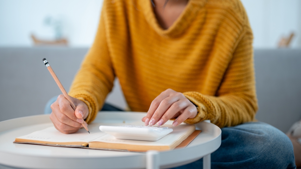 Mujer sentada en un sofá en casa, escribiendo en un libro y usando una calculadora para administrar sus gastos domésticos y calcular sus ahorros mensuales. personas y conceptos financieros