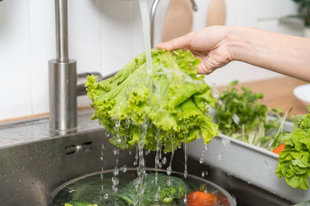 Cerrar la mano de una mujer joven asiática que lava el brote de girasol, lechuga de roble verde hidropónica, verdura fresca con agua de salpicadura en la cuenca del agua en el fregadero en la cocina en casa, preparando la ensalada, comida de cocina