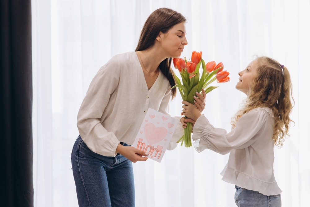 Niña alegremente presentando tulipanes rojos y una tarjeta de felicitación hecha a mano a su madre, celebrando el amor y el vínculo del Día de la Madre