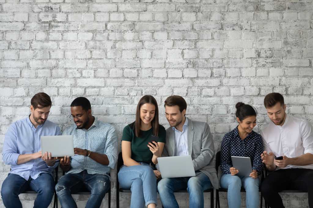 Cadena de información. Grupo de jóvenes multiétnicos diversos sentados en sillas por pares sosteniendo gadgets intercambiando datos en medios sociales, mostrando videos, descargando archivos 