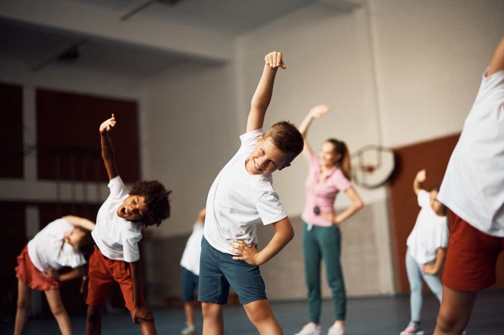 Grupo de estudiantes de primaria calentándose mientras hacen ejercicios de estiramiento en clase de educación física en el gimnasio de la escuela. El foco está en el chico feliz mirando la cámara. 