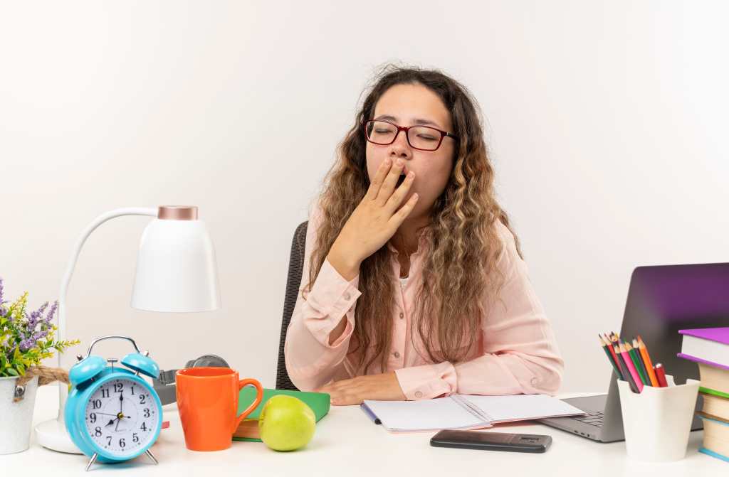 tired young pretty schoolgirl wearing glasses sitting at desk with school tools doing her homework putting hand on mouth and yawning with closed eyes isolated on white background