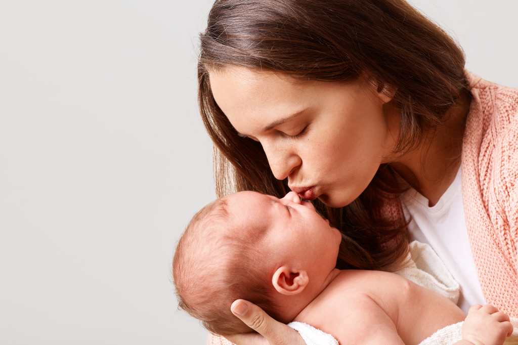 Closeup portrait of winsome woman kissing her newborn baby with closed eyes, expressing her love and care, lady in casual clothing holding her kid in hands, isolated over white background.