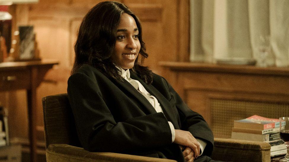 Ayo Edebiri, sentada sonriente con un traje negro en una escena de "After The Hunt". 