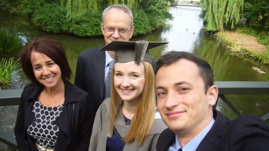 Jessica Brady viste una toga universitaria y un birrete, rodeada de su familia —su madre a la izquierda y su padre detrás— con un estanque y árboles de fondo.  