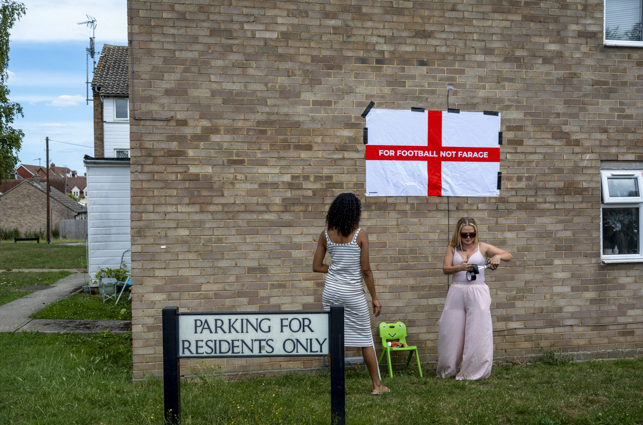 Flo Finch, right, tapes a customized message that reads "Football not Farage" across the red cross of an England national flag, known as the St. George's flag, outside her home in Essex, June 29, 2026. The national flag of England, a white banner with a bold red cross, has been displayed by soccer fans during the World Cup, but it has caused tension over whom, and what, it is meant to represent. (Andrew Testa/The New York Times)
